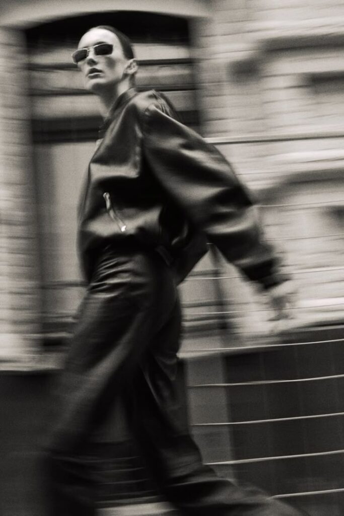 Black and white fashion photo of a model in motion wearing a leather jacket, leather pants, and sunglasses, walking through an urban setting with a blurred dynamic effect.