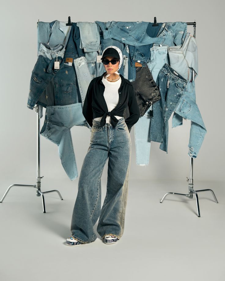 Fashion model wearing wide-leg jeans, a white top, black button-up shirt tied at the waist, sneakers, sunglasses, and a headscarf, standing in front of a backdrop of hanging denim pieces in various shades of blue.
